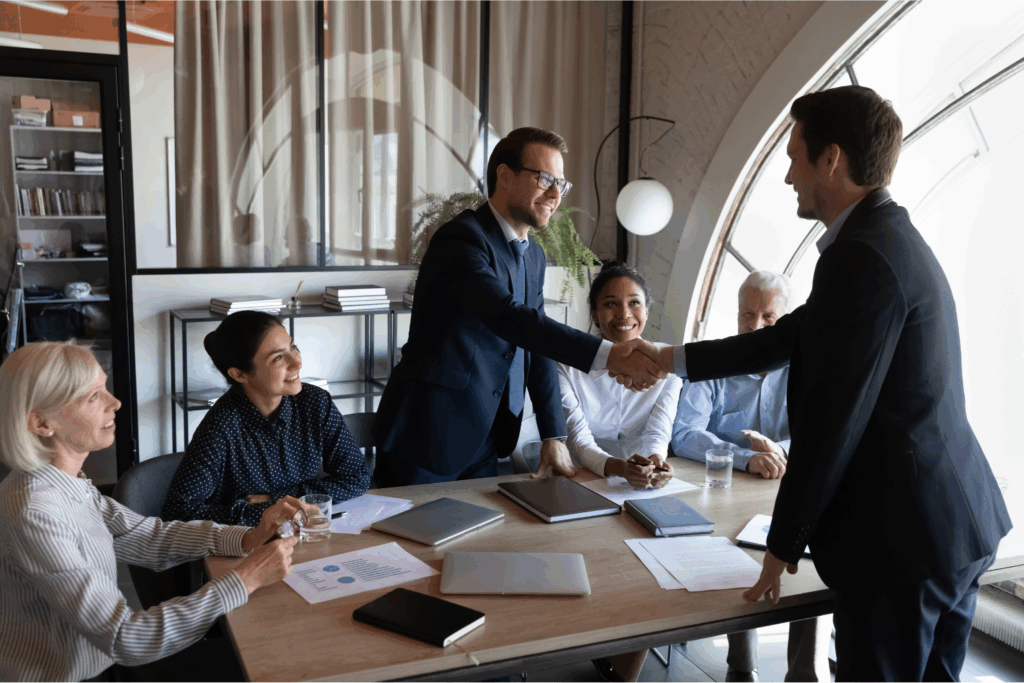 A diverse group of six business professionals sits around a modern office conference table. Two men in suits are standing and shaking hands across the table, smiling, while their colleagues observe the interaction with engaged and positive expressions. The scene takes place in a well-lit room with large arched windows and shelves in the background.