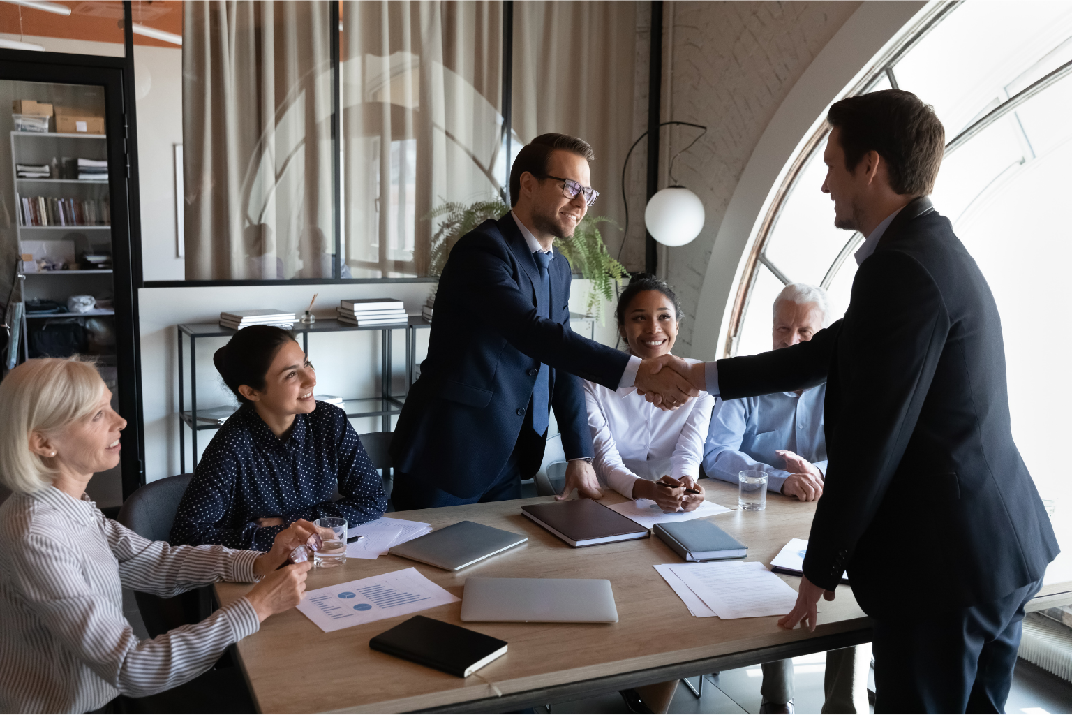 A diverse group of six business professionals sits around a modern office conference table. Two men in suits are standing and shaking hands across the table, smiling, while their colleagues observe the interaction with engaged and positive expressions. The scene takes place in a well-lit room with large arched windows and shelves in the background.