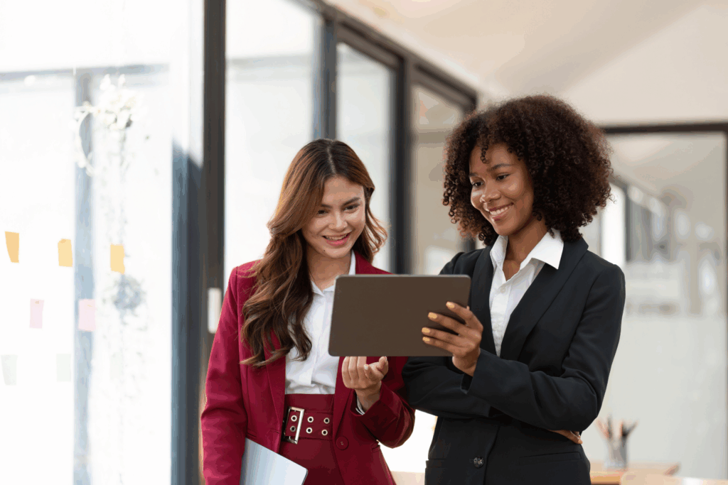 Two professional women in business attire stand together in a modern office, smiling and looking at a tablet. One woman, in a dark suit, holds the tablet while the other, in a red suit, holds a laptop and gestures toward the screen. Colorful sticky notes are visible on a glass wall in the background.