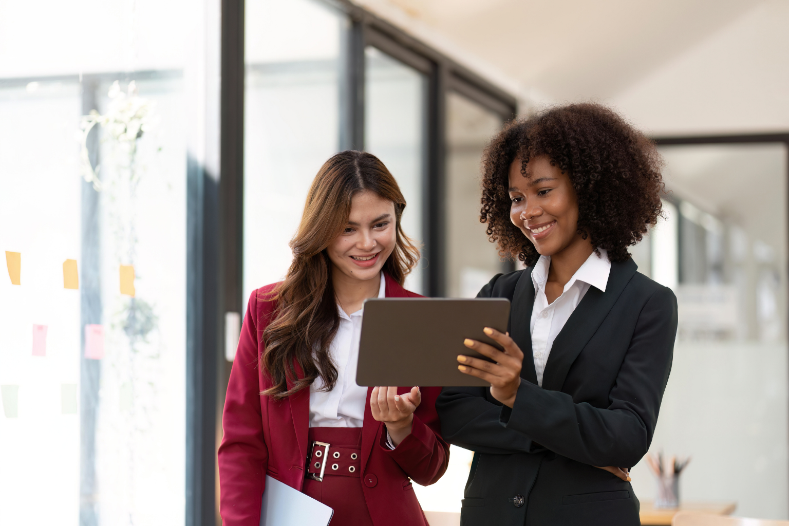 Two professional women in business attire stand together in a modern office, smiling and looking at a tablet. One woman, in a dark suit, holds the tablet while the other, in a red suit, holds a laptop and gestures toward the screen. Colorful sticky notes are visible on a glass wall in the background.